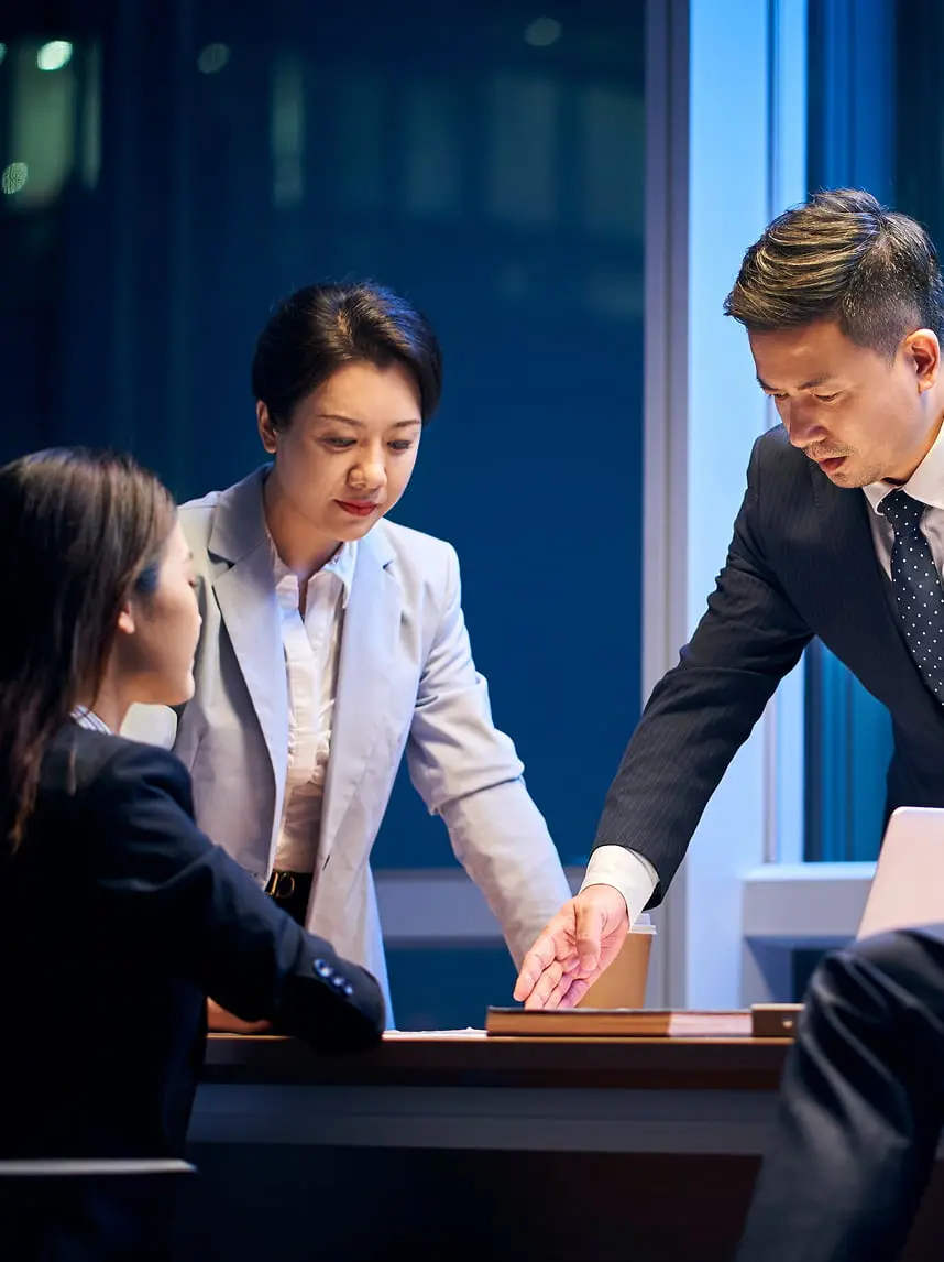 オフィスの会議室で3名のビジネスパーソンが書類を確認しながら打ち合わせをしている様子。夜のオフィスビルを背景に、スーツを着た男性と女性が机を囲んで真剣に議論している。プロフェッショナルな雰囲気の中で、チームワークとコミュニケーションを象徴的に表現している場面。